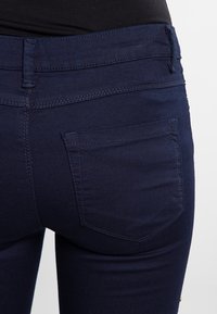 Jean en denim bleu marine au design ajusté, avec cinq poches et des coutures visibles sur la taille et les poches arrière.
