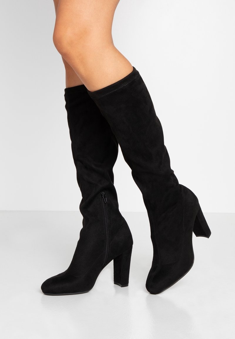 Botas negras por encima de la rodilla en suave ante, con un tacón grueso y un zipper lateral para el cierre. Textura suave y diseño ajustado.