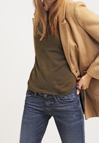 Abrigo de lana beige sobre un suéter verde oliva, combinado con unos jeans de mezclilla azul. Presenta texturas suaves y un diseño ajustado.