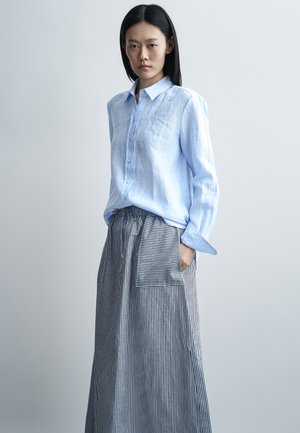 Tânără cu păr negru drept, purtând o cămașă albastru deschis cu nasturi și o fustă lungă cu dungi și buzunar mare în față.