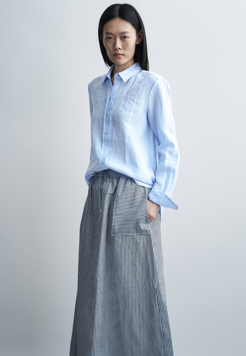Jeune femme aux cheveux noirs et raides portant une chemise bleue claire boutonnée et une longue jupe rayée avec une grande poche avant.