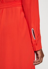 Abito rosso a maniche lunghe con una texture liscia, caratterizzato da una vita accentuata da una cintura e polsini con bottoni che presentano un dettaglio a righe contrastanti all'interno.