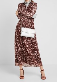 Long, sheer brown dress with white polka dots, buttoned collar, tied waist, paired with a silver clutch and brown heeled sandals.