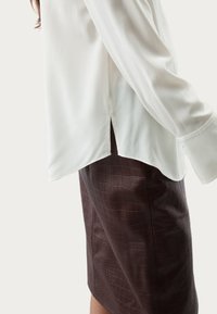 Blusa blanca de manga larga con una abertura lateral, combinada con una falda de cocodrilo texturada de color marrón oscuro. Tejido suave y corte moderno.