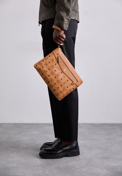Sac à main en cuir tan avec un motif embossé et une poche zippée à l'avant, tenu par une personne portant un pantalon noir et des chaussures noires.