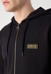 Black zip-up hoodie made of soft fabric, featuring a gold-accented logo patch, zipper, and drawstrings without additional embellishments.