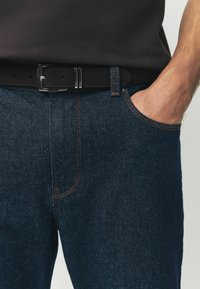 Jeans de mezclilla azul oscuro con costuras naranjas, que cuentan con un bolsillo frontal y un cinturón negro con una hebilla metálica brillante.