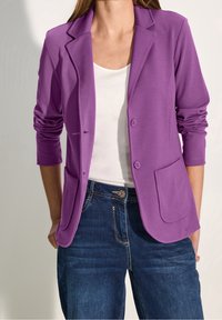 Purple, lightweight blazer with a notched collar, two front pockets, and two buttons. Paired with a white top and dark denim jeans.