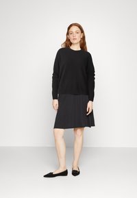 Mustra mustre, mille pikkade varrukatega, musta plisseeritud kleidi peal. Mudel kannab musti madalaid kingi, millel on kerge sära. Lihtne, minimalistlik disain.
