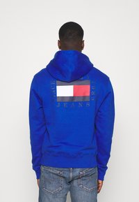 Kungsblå luvtröja med ett grafiskt tryck som har en rektangulär flaggdesign i rött, vitt och blått på baksidan.