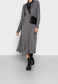 Femme portant une robe grise à manches longues, des bottines noires à la cheville et un sac bandoulière noir, marchant devant un fond blanc uni.