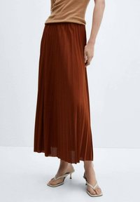 Mujer vestida con una falda midi plisada de color óxido, una blusa sin mangas de punto beige y sandalias de tacón beige, de pie frente a un fondo blanco liso.