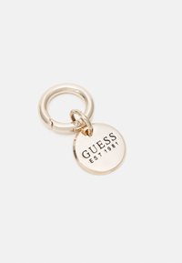 Porte-clés doré avec un pendentif rond gravé de "GUESS EST 1981" sur un fond blanc uni.