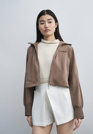 Młoda kobieta ubrana w krótki brązowy sweter na zamek błyskawiczny na beżowej bluzce i białe plisowane spodenki z wysokim stanem na jednolitym szarym tle.