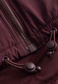 Burgundi színű dzseki részlete, YKK cipzárral, húzózsinórral és kapcsolókkal, valamint egy finom gyémánt mintázatú textúrával.