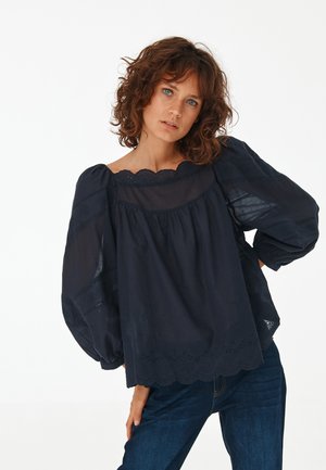 Femme aux cheveux châtain bouclés et aux yeux bleus, portant un chemisier ample bleu foncé à manches bouffantes et un jean bleu, debout devant un fond uni.