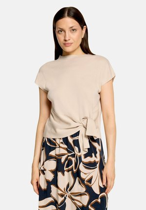 Femme aux cheveux raides et foncés portant un haut beige à manches courtes noué à la taille et une jupe bleu marine avec de grands motifs floraux beige et marron.