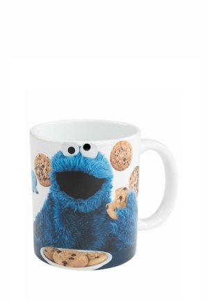 Weiße Tasse mit blauem pelzigem Charakter, der Schokoladenkekse hält und von ihnen umgeben ist, mit einem Teller Kekse davor.