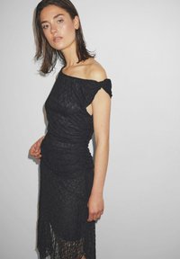 Fekete csipkés ruha, váll nélküli fazonnal, ráncolással a deréknál és egy középhosszú, áttetsző szegéllyel, amely extra textúrát és részleteket ad.