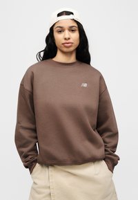 Καφέ φούτερ με λαιμόκοψη τύπου crew και ριμπ μανσέτες, με μικρό κεντημένο λογότυπο στο στήθος. Συνδυάζεται με μπεζ παντελόνι.