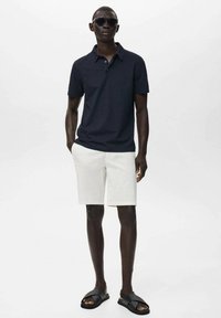 Námořnické polo tričko, bílé šortky a černé sandály. Sluneční brýle a krátké rukávy vytvářejí ležérní outfit. Hladká textura a přiléhavý střih.