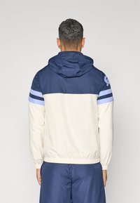 Hooded jack in marineblauw en crème, met blauwe strepen op de mouwen en elastische manchetten. Beschikt over een gladde, lichte stofstructuur.