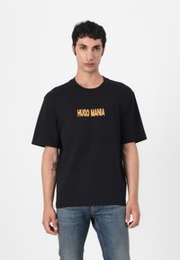 Svart bomull T-skjorte med "HUGO MANIA" trykket i oransje tekst. Korte ermer, rund hals, avslappet passform. Brukt sammen med blå jeans.