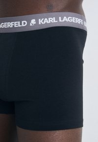 Svart boxershorts med en grå midjeband der "KARL LAGERFELD" er trykt i hvitt. Glatt stoff, tettsittende passform, midt på låret.