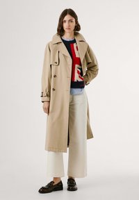 Beige trenchcoat med klassisk krage, dubbelknäppt design och bälte vid ärmsluten, över en tröja med kraftiga röda och blå mönster.