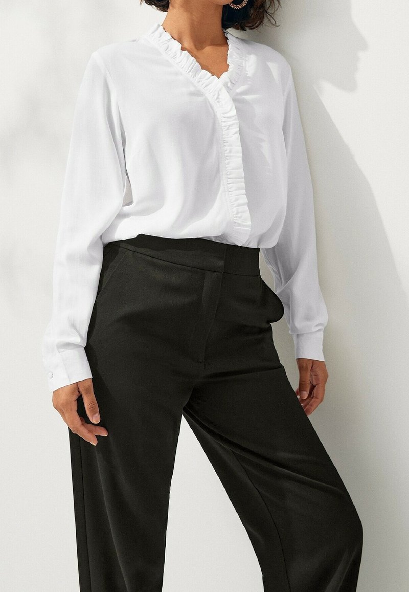 Femme portant un chemisier blanc à manches longues avec un col en V à volants et un pantalon large taille haute noir, debout contre un mur blanc.