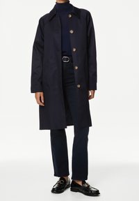 Persona vestida con un abrigo botoné navy encima de un cuello de tortuga oscuro, vaqueros oscuros con un cinturón negro y mocasines de cuero negros con hebillas doradas.