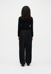 Γυναίκα με σγουρά μαλλιά που στέκεται με την πλάτη, φοράει μαύρο μακρυμάνικο τοπ και άνετο, πιετέ μαύρο παντελόνι σε απλό φόντο.