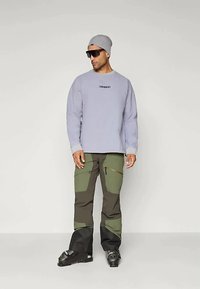 Ανοιχτό μωβ φούτερ με λογότυπο, σκούρα πράσινα και μαύρα cargo παντελόνια με τσέπες και φερμουάρ, γκρι σκουφί και γυαλιά ηλίου.