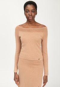 Off-shoulder ribbet top i lys beige, med lange ærmer og en tætsiddende silhuet, samt en lille guldlogo detalje i kanten.