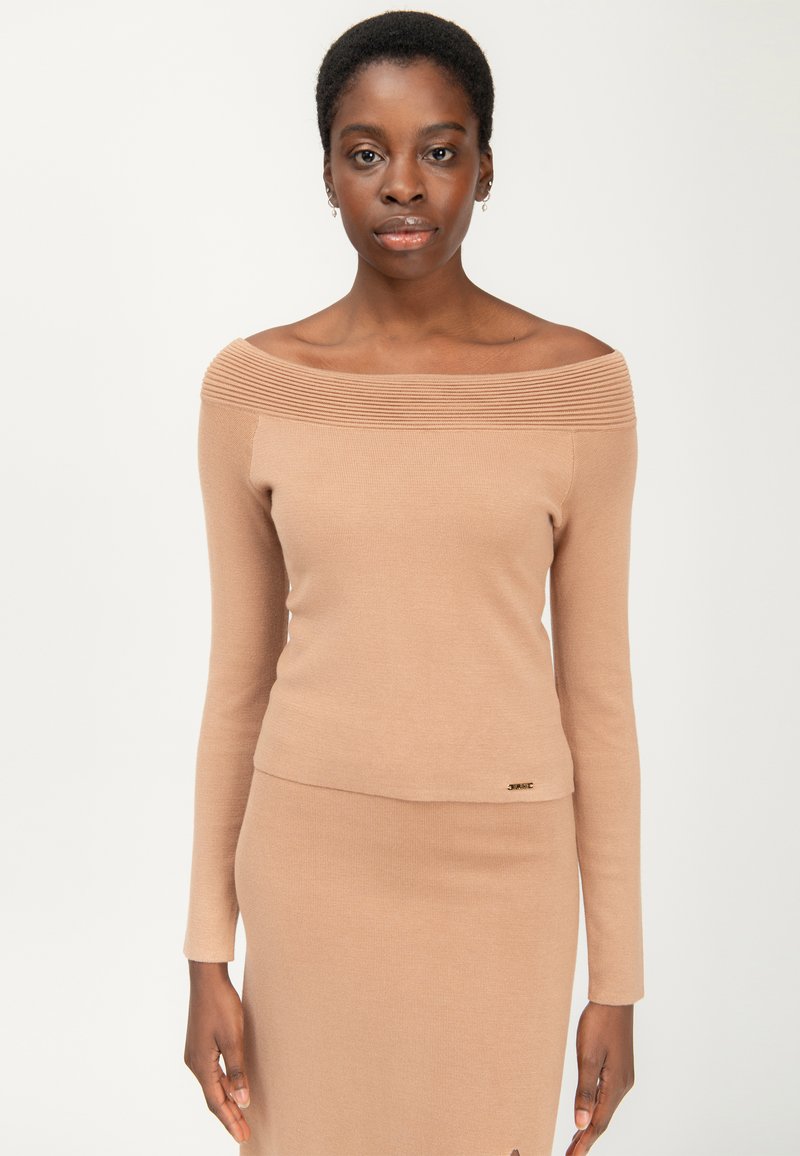 Off-shoulder ribbet top i lys beige, med lange ærmer og en tætsiddende silhuet, samt en lille guldlogo detalje i kanten.