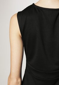 Haut sans manches noir en tissu lisse et extensible, avec une taille cintrée et un col rond. La vue de dos présente des lignes épurées et un minimum de détails.