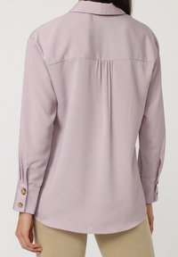 Blusa de manga larga color lila con cuello puntiagudo, pliegue en la espalda y puños abotonados. Tela suave con un ligero brillo.