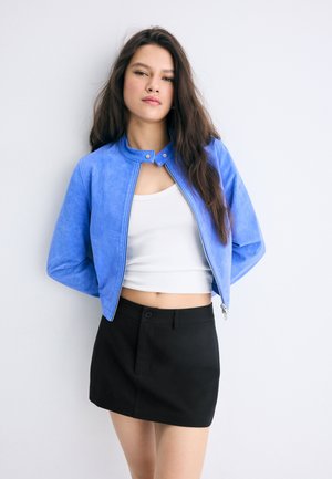 Jeune femme aux longs cheveux foncés portant une veste bleue, un crop top blanc et une jupe noire, debout sur un fond blanc.