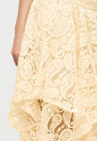 Gros plan sur une main à côté d'une jupe en dentelle crème avec des motifs paisley et floraux superposés sur un tissu transparent.