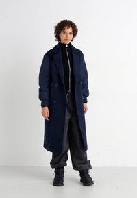 Navy blue long coat with a double-breasted design, wide sleeves, and a smooth texture, worn over a black ribbed top and gray pants.