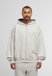 Hombre con barba corta que lleva puesta una sudadera con cremallera de color gris claro y pantalones de chándal a juego, de pie frente a un fondo liso de color gris claro.