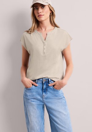 Femme portant un haut beige à manches courtes boutonné, un jean bleu clair, une casquette beige à motifs et des boucles d'oreilles créoles dorées, debout les mains dans les poches.