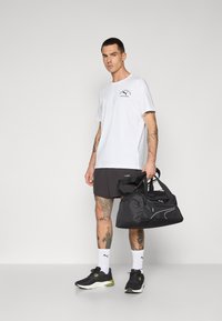 Hvid t-shirt med logo, sorte shorts med brandnavn, sorte sportsko og sort duffelbag med hvide accenter. Glat tekstur.