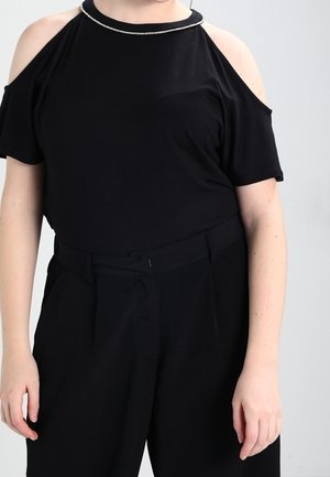 Osoba nosząca czarną bluzkę z odkrytymi ramionami z ozdobnym dekoltem i czarne spodnie z wysokim stanem na jasnym tle.