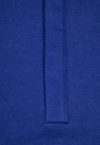 Gros plan sur un tissu bleu, avec une couture verticale et un rabat rectangulaire renforcé, montrant la texture et les détails de la couture.