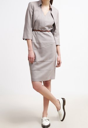 Kvinde iført en lysegrå knælængdekjole med brun bælte, matchende kort blazer og hvide slip-on sko med sorte såler.