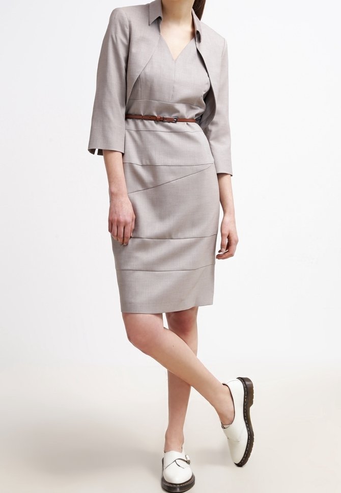 Femme portant une robe mi-longue gris clair avec une ceinture marron, un blazer court assorti, et des chaussures blanches à enfiler avec des semelles noires.