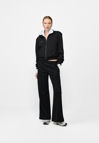 Czarna kurtka zapinana na zamek z białym kołnierzem, zestawiona z luźnymi czarnymi spodniami. Oba elementy mają gładką teksturę i minimalne detale brandingowe.