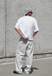 Bela oversize majica, svetlo sivi široki hlače, sivi kapu i crne cipele, stojeći uz teksturirani sivi zid, ruku iza leđa.