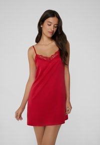 Abito sottoveste in satin rosso con bordi in pizzo lungo il collo. Presenta sottili spalline e una silhouette fluida e liscia.